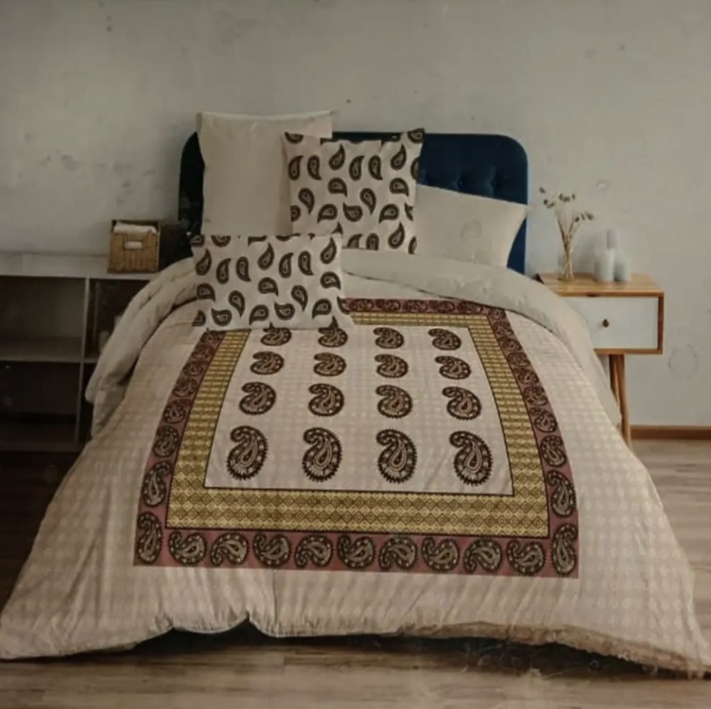 Beige paisley printed cotton bedspread with maroon border and matching cushion covers on blue upholstered bed